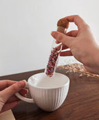 Uma pessoa segura um infusor de vidro acima de uma xícara de chá fumegante, pronto para infundir a bebida.